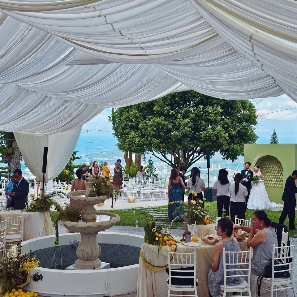 A wedding reception at Hillside Kokol under a white draped canopy, with a stone fountain, round tables and the sea visible past the trees